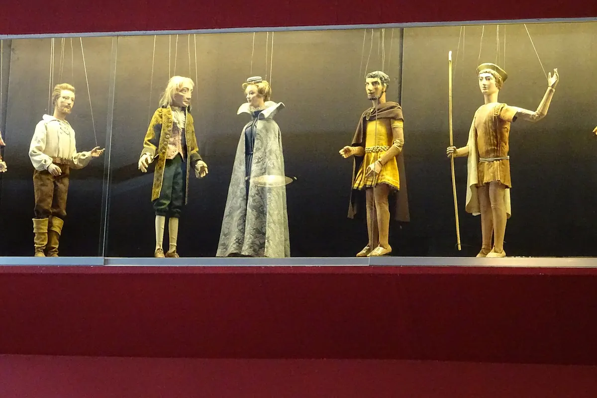 Five marionettes in period costume hang from strings inside a lit glass case against a deep red wall