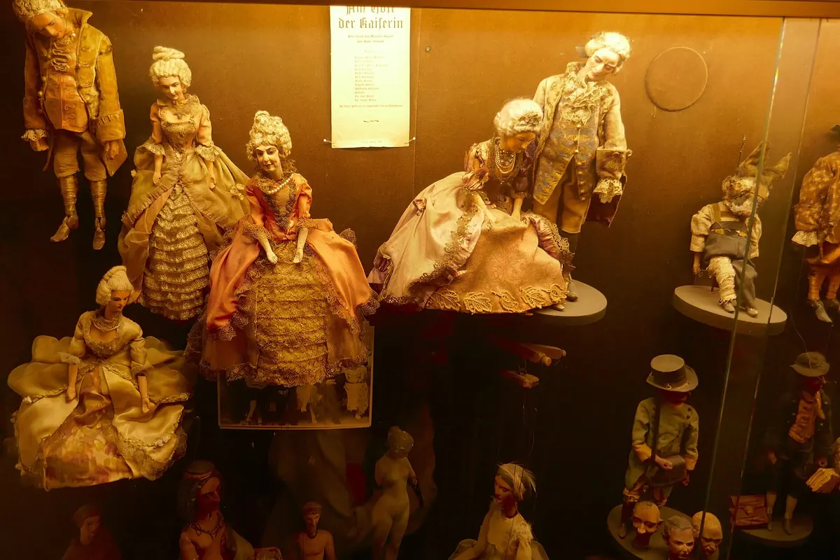 Marionettes in eighteenth-century dress with powdered hair and full skirts stand inside a lit display case