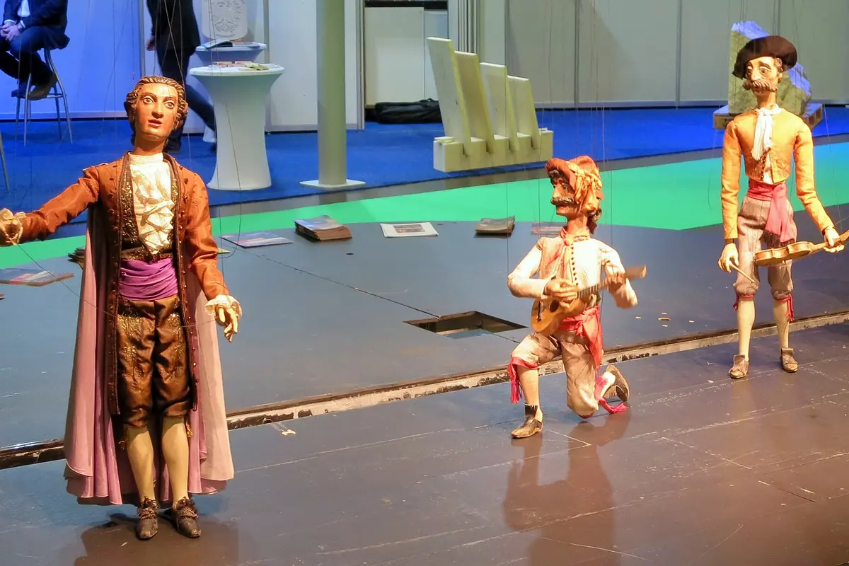 Three marionettes in eighteenth-century costume stand on a bare stage: a tall figure in a purple-lined cloak, a kneeling figure with a lute and a standing figure with a hat